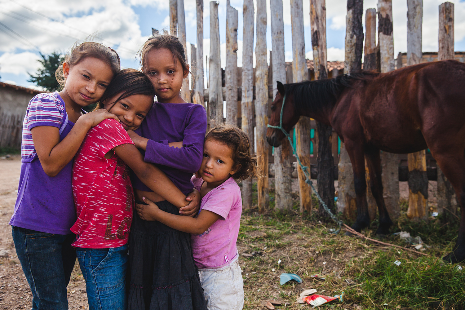 Four young girls hold one another looking at the camera with a horse in front of a fence in the background.