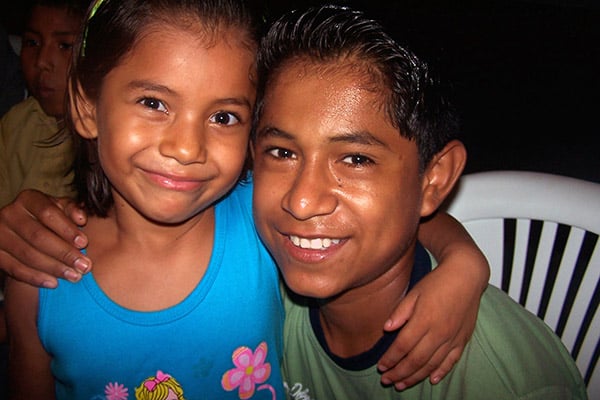 NPH Bolivia Young girl with dark shoulder length hair and an older boy with their arms around one another smiling at the camera.