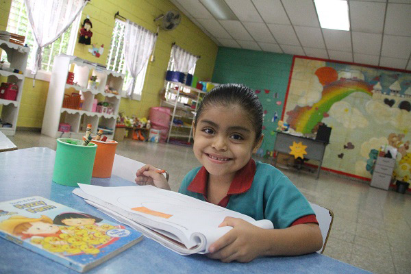 NPH El Salvador_Niña con el cabello recogido, sonriendo sobre un cuaderno de ejercicios blanco, sosteniendo un lápiz
