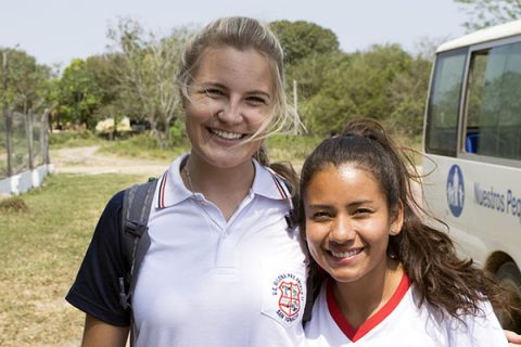 NPH Bolivia Volunteer_Smiling blond young woman in a white polo standing alongside a younger smiling brunette wearing a white shirt with a red collar in front of a white bus